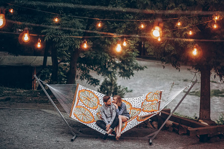 时尚时髦的情侣拥抱和放松在吊床下的复古灯在傍晚的夏季公园。顶部视图 