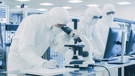 tt防护摄影照片_在《穿着防护服做研究的实验室科学家》中，使用显微镜并将数据写入笔记本。现代手工生产半导体和药品.