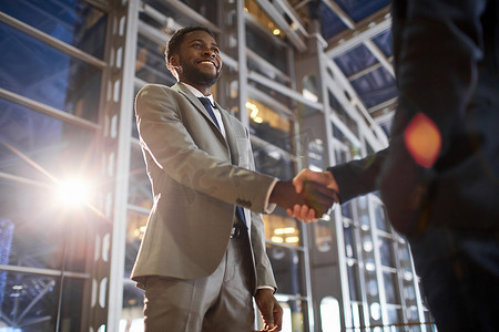 两个商业伙伴握手时的低角度肖像: 微笑的非洲裔美国商人在现代玻璃办公楼大厅与白种同事握手