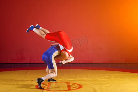 红蓝圆框摄影照片_两名身穿红蓝相间制服的男子摔跤选手在体育馆的一块黄色摔跤地毯上摔跤