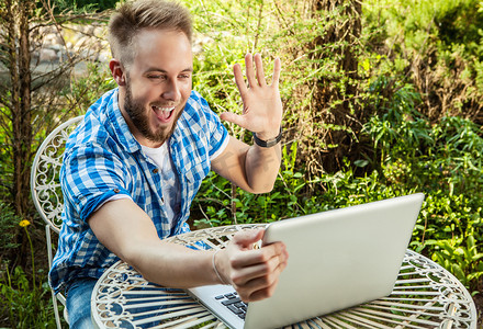 Young smiling handsome man in casual clothes work at an iron table with computer against country garden.