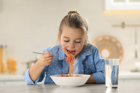 饭桌摄影照片_可爱的小女孩在厨房的饭桌上吃着美味的意大利面