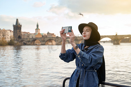 bridge摄影照片_Stylish young beautiful woman earing black hat in Prague with Charles Bridge on background. Elegant retro lady fine art portrait.
