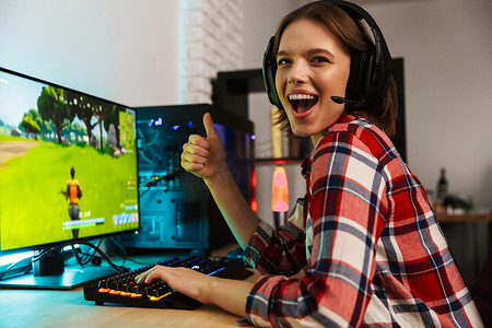 一个兴奋的年轻女子戴着耳机坐在桌旁, 玩电脑网络游戏的肖像
