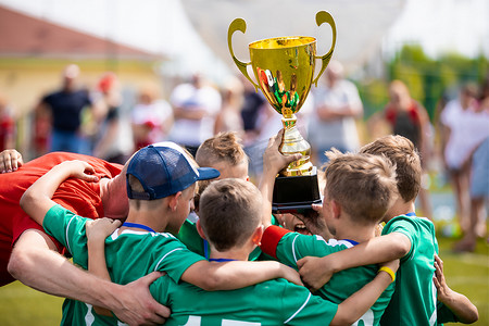 多色奖杯印章摄影照片_抱着奖杯的年轻足球运动员。男孩们庆祝足球足球冠军。为孩子儿童赢得团队的体育比赛.