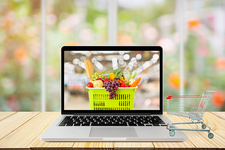 laptop摄影照片_laptop computer and shopping cart on wood table with window and garden abstract blur background grocery online concept