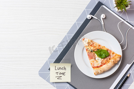 在白色木桌上刻有午餐时间的比萨饼、耳机和便条的顶视图