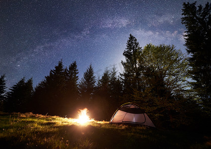 晚上露营在山上。旅游帐篷由明亮的燃烧篝火附近的森林在清澈的深蓝色星空, 银河。背景高的松树。自然美与旅游理念.