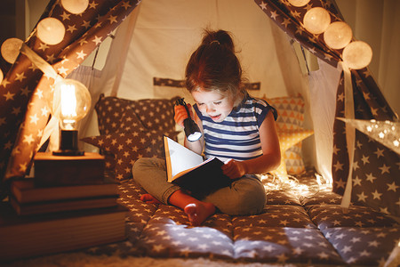 哈哈大笑卡通人物摄影照片_快乐的儿童女孩在黑暗的帐篷里大笑看书