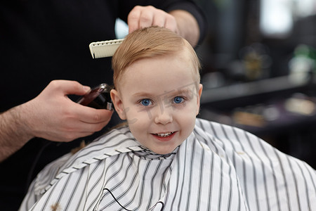 可爱的金发碧眼的微笑的男婴与蓝色的眼睛在理发店有理发由美发师。有工具的造型师之手。儿童时尚。室内, 黑暗的背景, 复制空间.