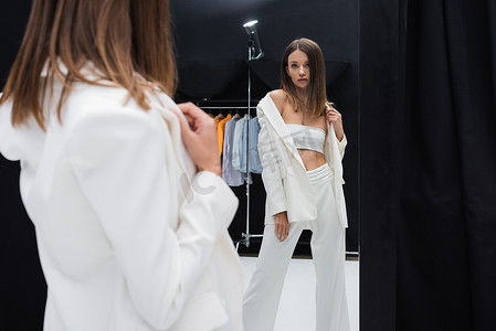 brunette model in white suit posing near mirror in photo studio