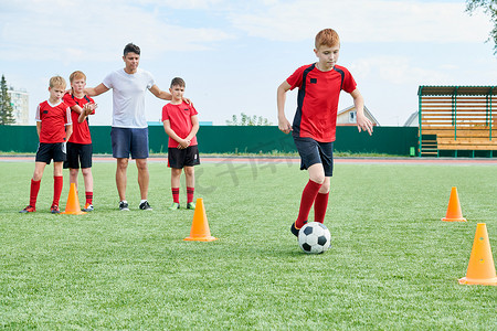 全长肖像的初级足球队训练户外在阳光下, 重点是红头发男孩领先的橙色锥之间的球, 复制空间