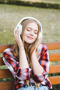 穿着休闲服装的美丽女孩的肖像照片，坐在公园的木凳上，用闭着眼睛在耳机上沉思