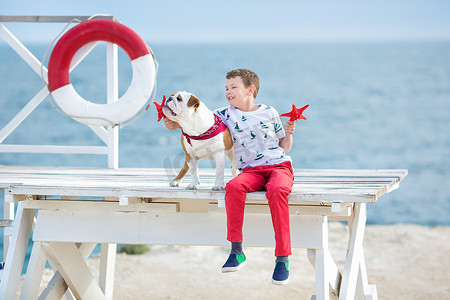 英俊的男孩青少年 happyly 花时间与他的朋友斗牛犬在海边的孩子狗控股玩两个海星接近生活浮标穿红色长裤拖鞋和 t-shirt