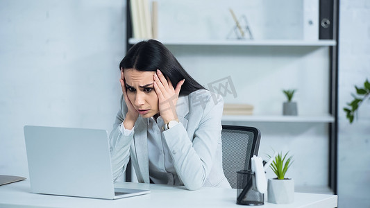 stressed businesswoman touching head and looking at laptop on desk