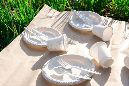 可生物降解的一次性餐具，由环保材料制成。牛皮纸上的汤匙、叉子、玻璃杯和天然玉米淀粉盘。草地上的阴影。特写镜头
