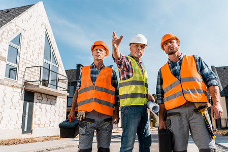 戴钢盔摄影照片_戴钢盔的建筑工人在靠近房屋的地方拿着工具箱 