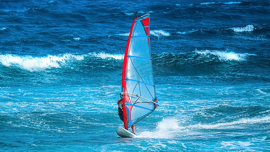 红蓝圆框摄影照片_夏季体育运动：在一个多风的夏日下午，一个身披红蓝帆的冲浪运动员在波浪中冲浪.
