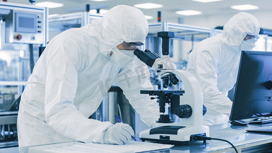 tt防护摄影照片_在实验室，穿着防护服的科学家利用显微镜进行研究，并将数据输入个人计算机。现代手工生产半导体和药品.