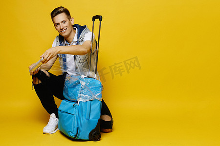 旅行箱广告摄影照片_一个穿着斜纹棉布衣服的年轻人正在准备把他的旅行箱放进行李箱里，这个行李箱被黄色的背景隔开了。旅游广告的调侃