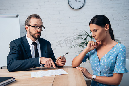 戴眼镜、留着胡子的律师一边拿笔靠近女职员一边用手指指着保险合同 