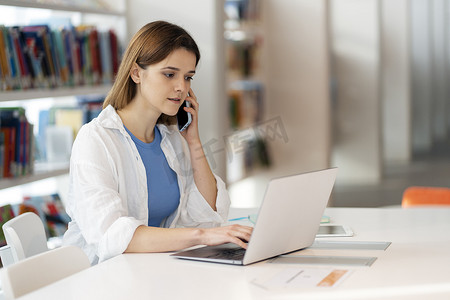 Young pensive woman talking on mobile phone and working on laptop online in modern office. Serious freelancer sitting at media library 