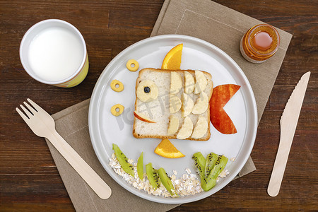有趣可爱的鱼形状三明治烤面包与香蕉，苹果，橙子，牛奶，蜂蜜。孩子们的甜甜点健康的早餐午餐食品艺术盘中，木制背景近景，俯瞰.