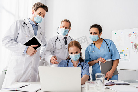 戴口罩的多族裔医生和护士使用手提电脑，同时在模糊的前景文件附近工作 