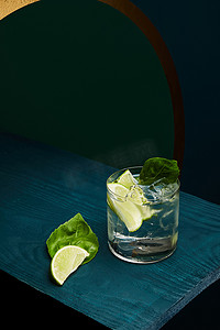 好看的几何形摄影照片_带有新鲜饮料、薄荷叶和石灰片的老式玻璃在蓝色木制表面绿色和蓝色几何背景的高角形视图