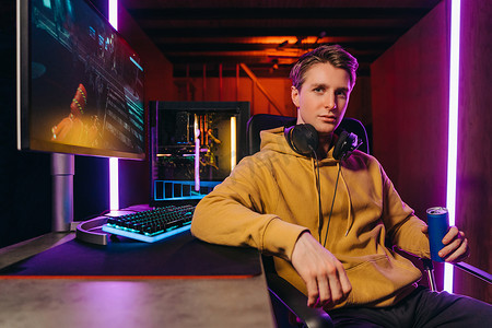 Portrait of young serious handsome pro gamer sitting at desk with gaming equipment, holding energy drink, using powerful PC for playing online video shooter games. Neon Colored Room, e-Sport concept 