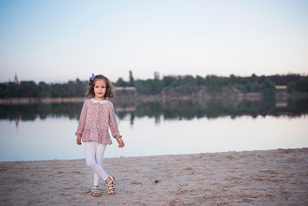 在湖边白色长裤的时髦女孩。孩子在沙子上赤脚花钱。美丽的小女孩在水库附近摆姿势。