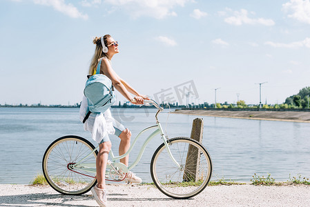 侧视图的女孩与背包骑自行车和听音乐在耳机附近的河在夏天