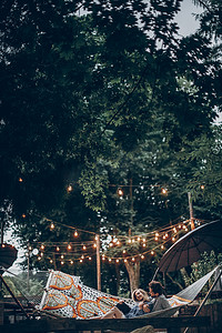 时尚时髦情侣拥抱和放松在吊床下的复古灯在傍晚夏季公园. 