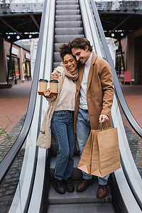 在商场里，一对快乐的多种族夫妇抱着纸杯和购物袋坐在扶梯上 