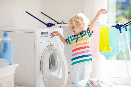 儿童在洗衣房内设有洗衣机或滚筒烘干机。帮助家庭琐事的孩子。现代家庭设备和洗涤洗涤剂在白色阳光的家。烘干机架清洗洗衣服. 