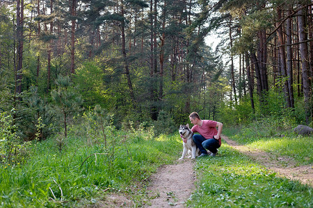 有魅力的年轻人穿着粉红色的 t恤衫和蓝色牛仔裤, 他的狗阿拉斯加在森林里散步。