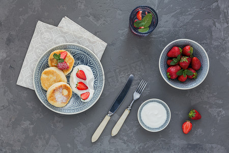 在灰色背景下, 早餐或午餐用酸奶奶油和草莓做的干酪煎饼。健康食品概念。平躺