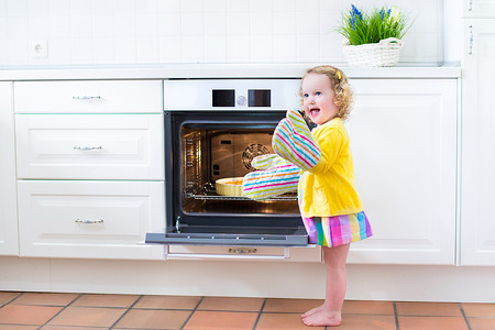 pi摄影照片_卷曲的蹒跚学步女孩在厨房旁边与苹果 pi 烤箱手套