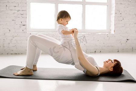 运动会幕布摄影照片_年轻快乐的妈妈锻炼身体，做臀部桥运动，穿着白色运动服，肚子上的小女孩，健康，产后瑜伽。 健康生活方式概念