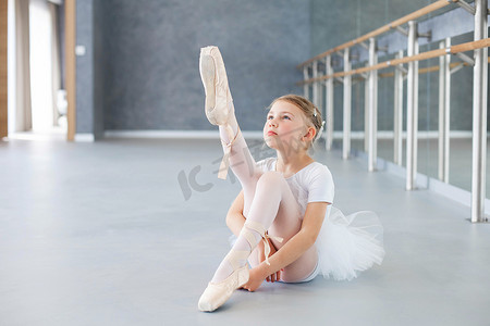 芭蕾班里的小芭蕾舞演员正在试穿尖皮鞋. 可爱的小女孩坐在理发店下面的地板上。 那孩子穿着白色芭蕾服装，穿着迷你裙跳舞.