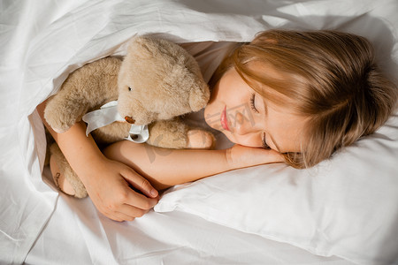 抱着泰迪熊的女孩睡在床上，床上铺着白布