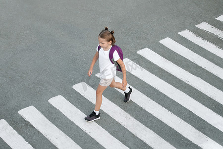 女学生在去学校的路上过马路。斑马的交通走在城市的路上。过人行横道的概念行人。时髦的少女背着背包走着。活跃的孩子。顶部视图