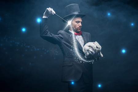 魔术师摄影照片_穿着西服、头戴帽子的情绪魔术师，带着魔杖和白兔表演魔术，黑漆漆的房间里挂满了烟雾和闪光的插图