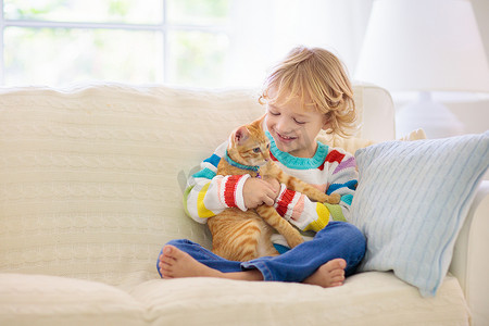 小孩和猫玩。抱着小猫的小孩小男孩抱着可爱的宠物坐在家里阳光灿烂的客厅里的沙发上。孩子们和宠物玩耍。儿童和家畜.