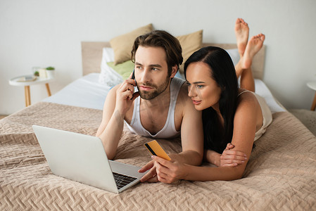 man holding credit card and talking on smartphone near laptop while shopping online with girlfriend 