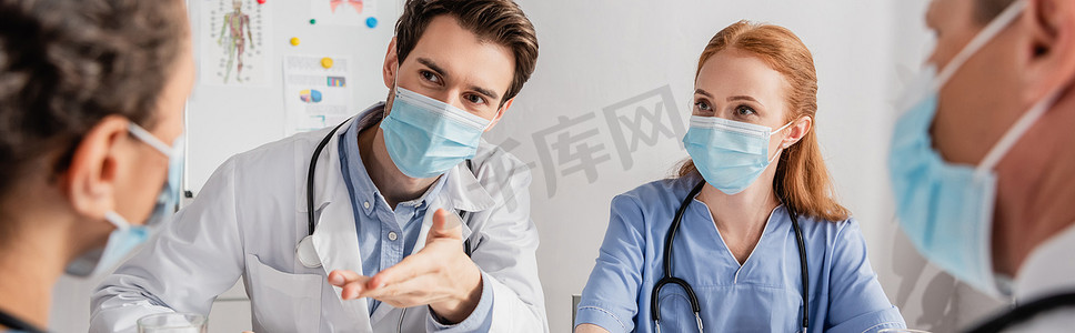 戴着医疗面罩的医生坐在同事身边，手拉手对着医院里的非洲裔美国护士，对着模糊的前景、横幅