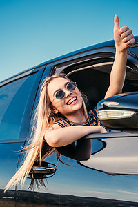 微笑时尚的妇女在太阳镜倾斜从汽车窗口和做拇指向上手势