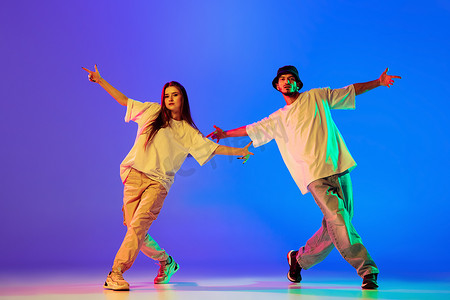 嘻哈女摄影照片_两个年轻人，穿着休闲装，跳着当代舞的男孩和女孩，在霓虹灯下跳着蓝底嘻哈.