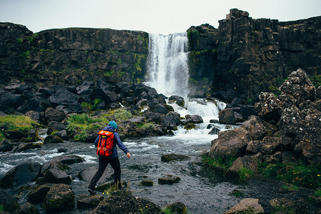 冰岛瀑布前中年妇女背着背包踩在河里岩石上的背影