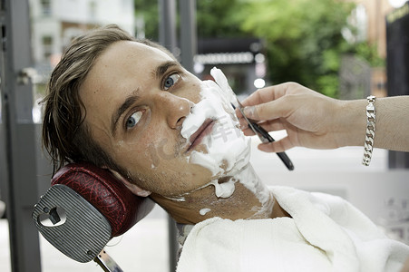 理发店里的年轻人脸上涂着剃须膏用剃须刀刮脸目光转向别处